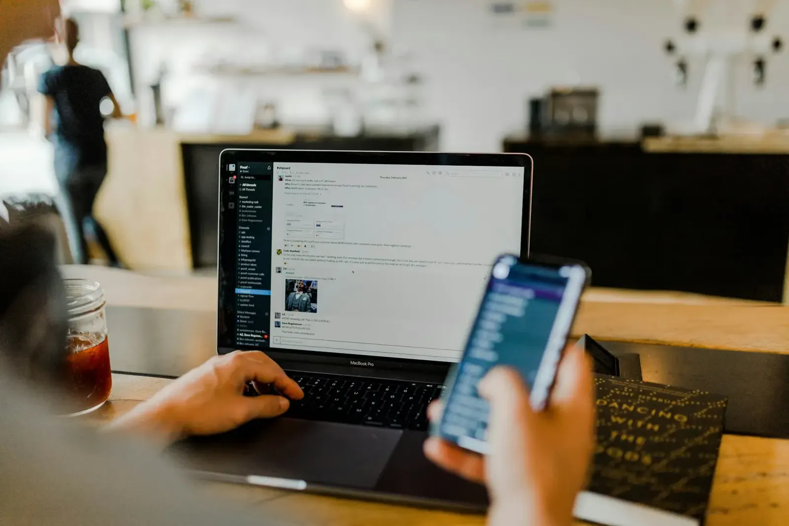 Person working on a laptop while checking a smartphone at a wooden desk